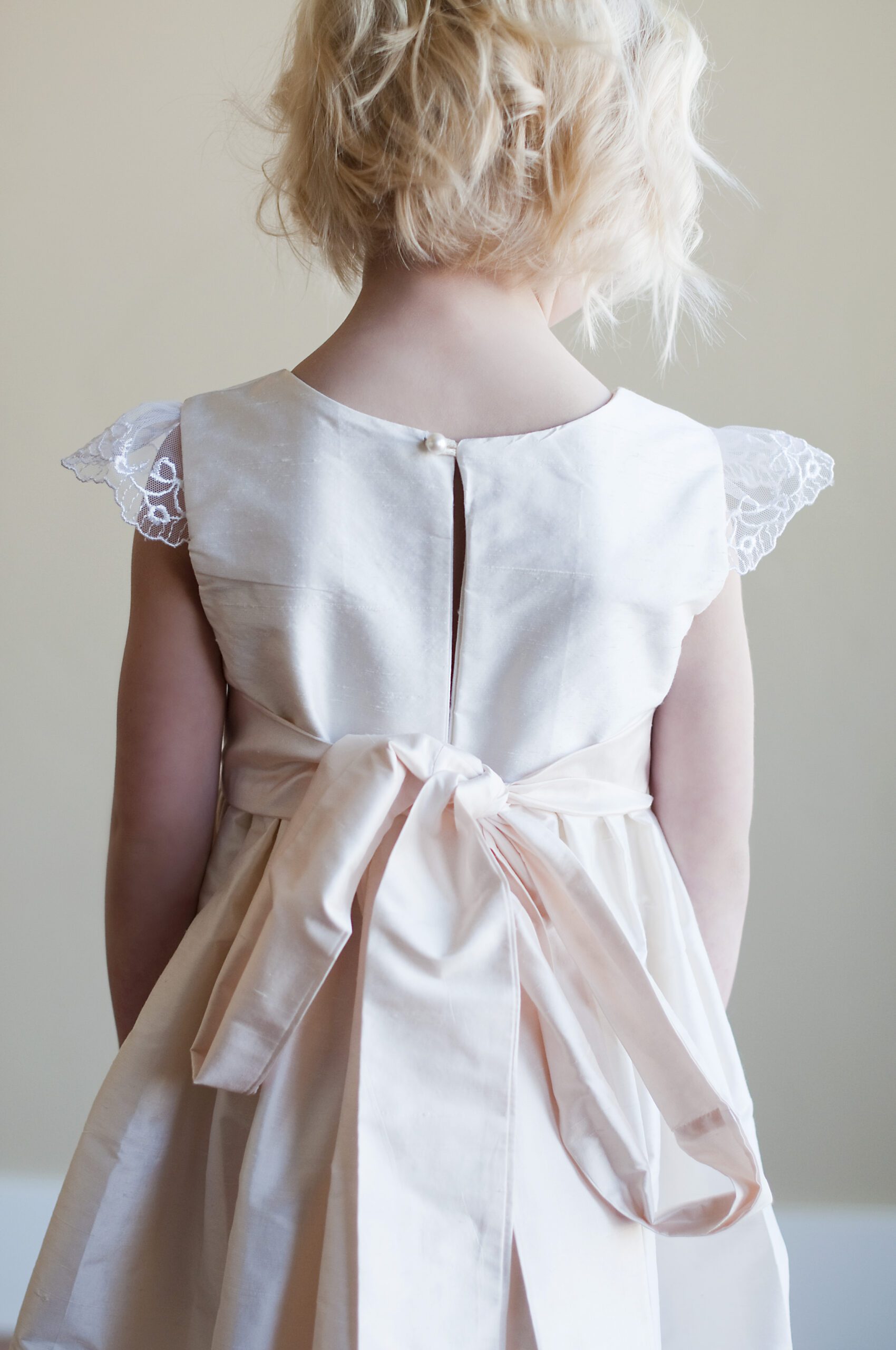 A photo of a pure silk flower girl dress in blush pink with lace sleeves and a big bow