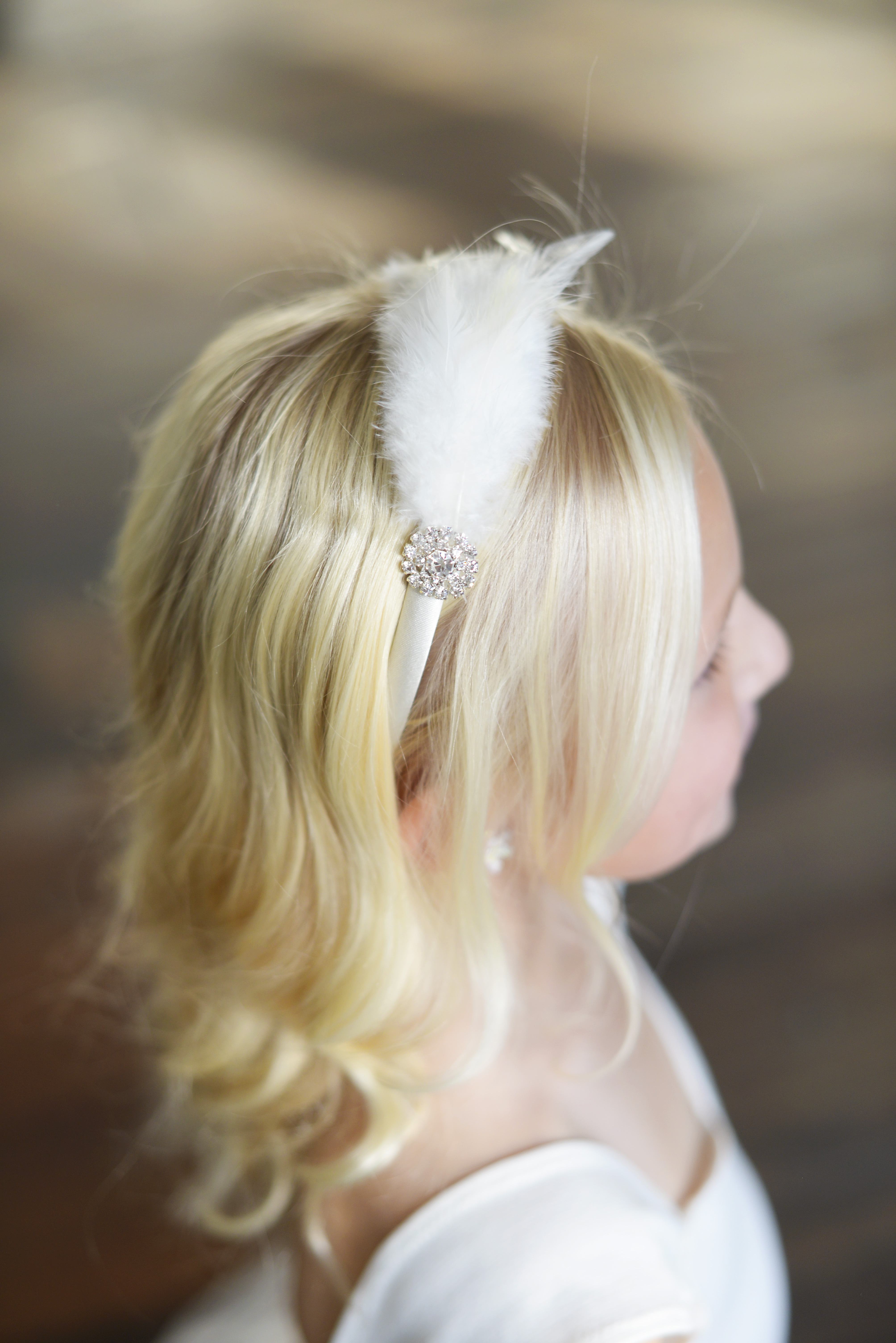 A photo of a flower girl wearing an ivory or white satin headband with a feather and diamante trim