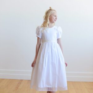 A photo of an ivory or white cotton first communion dress with a lace sash