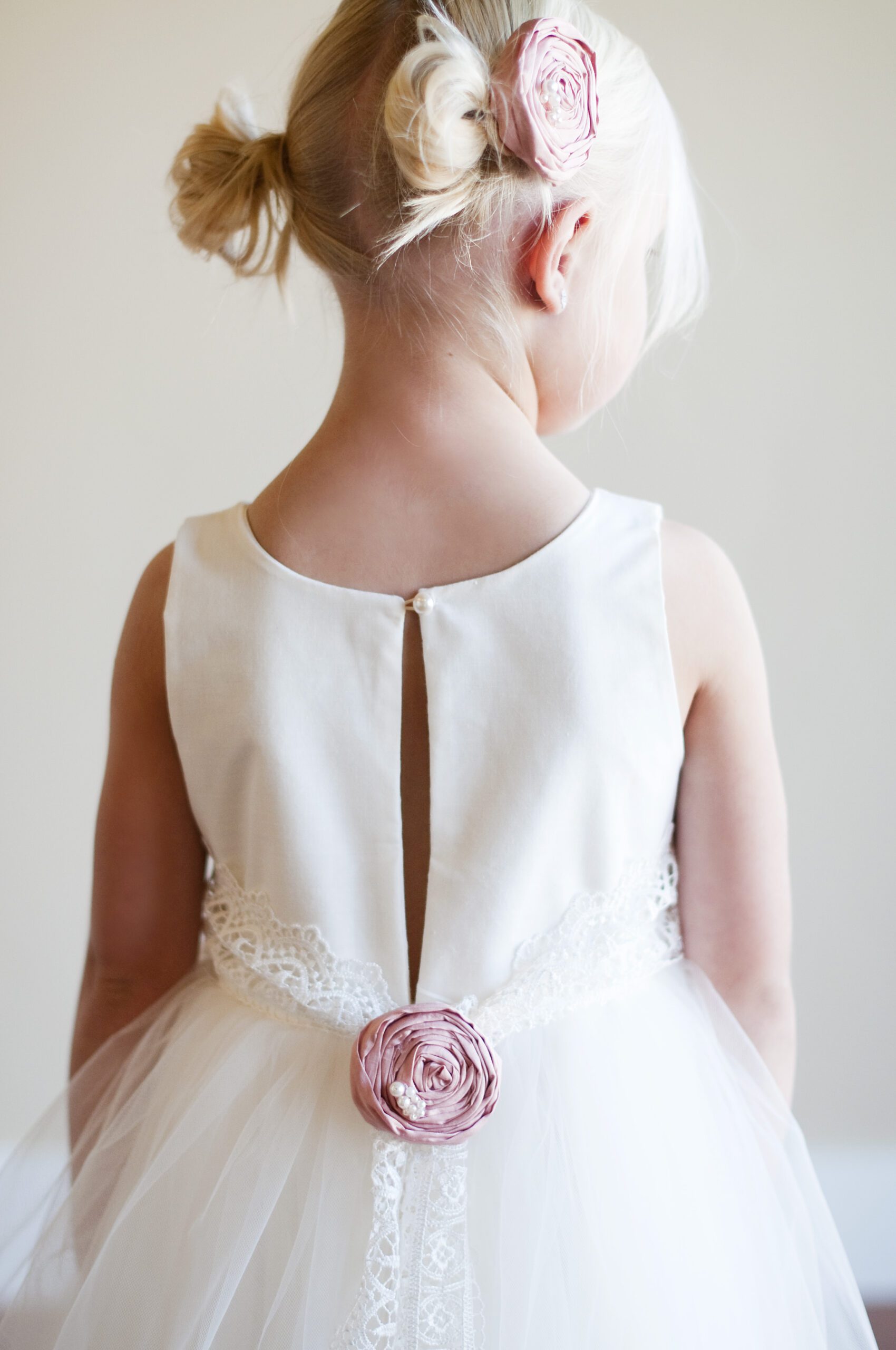 A photo of a flower girl wearing an ivory cotton dress with a tulle skirt and a lace belt. The belt has a blush pink silk rose.