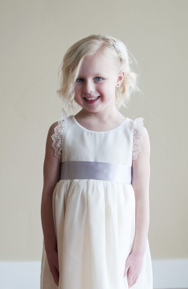A photo of a three year old girl with blond hair wearing an ivory chiffon flower girl dress with a silver sash and lace sleeves