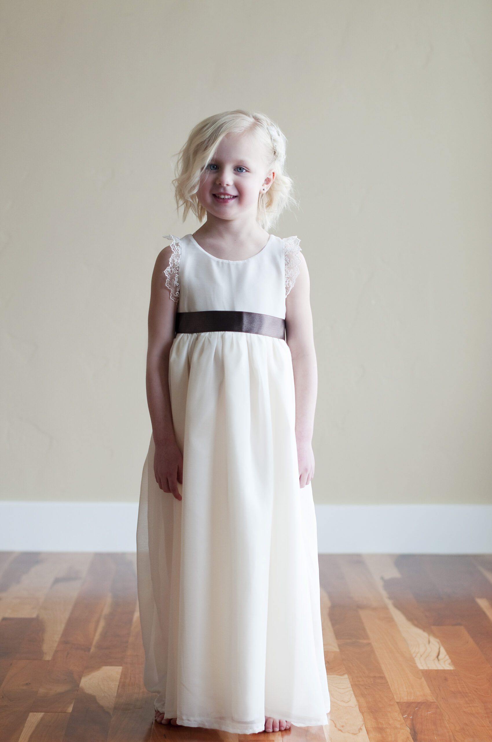 A photo of a three year old girl with blond hair wearing an ivory chiffon flower girl dress with a silver sash