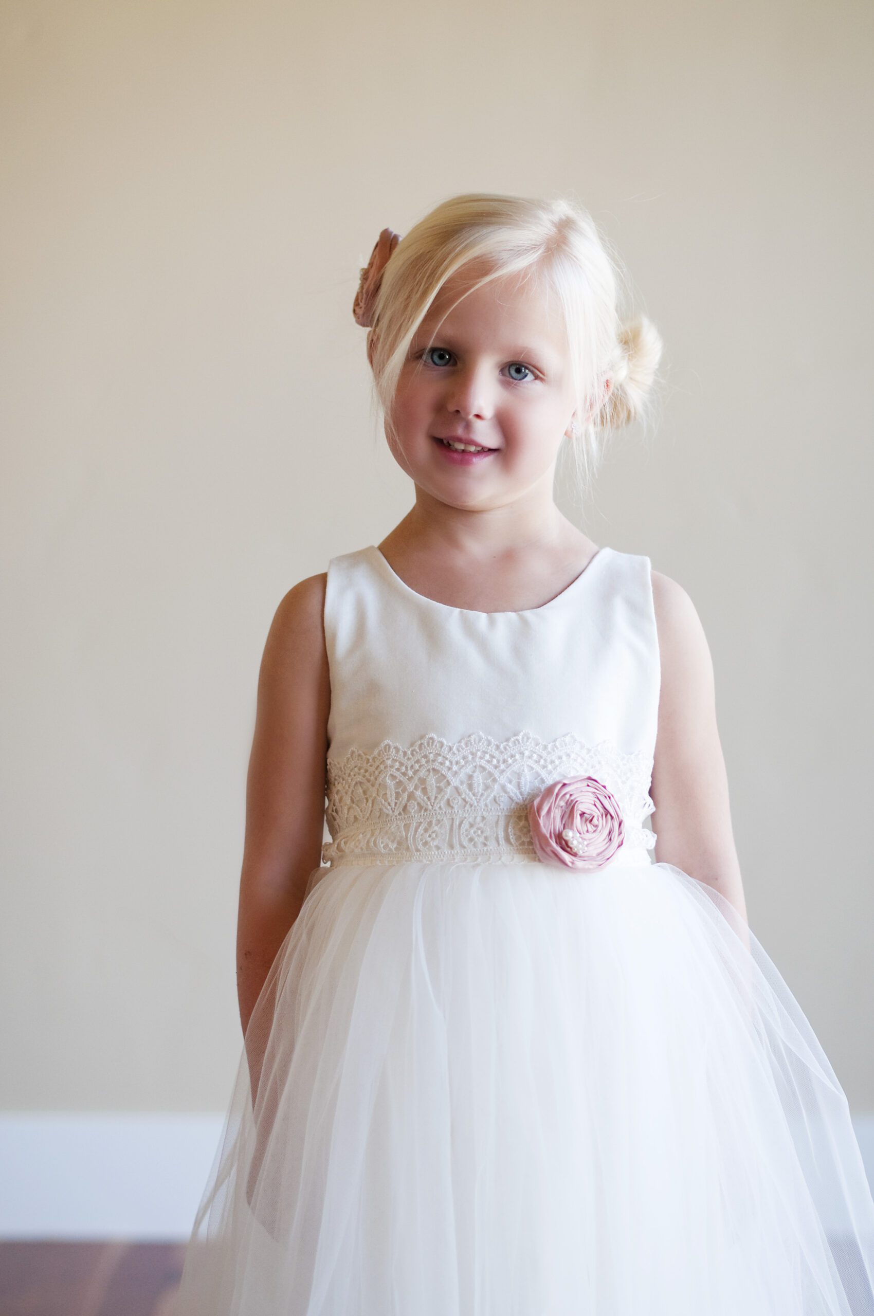A photo of a young flower girl wearing a white cotton and tulle flower girl dress with a pink silk flower