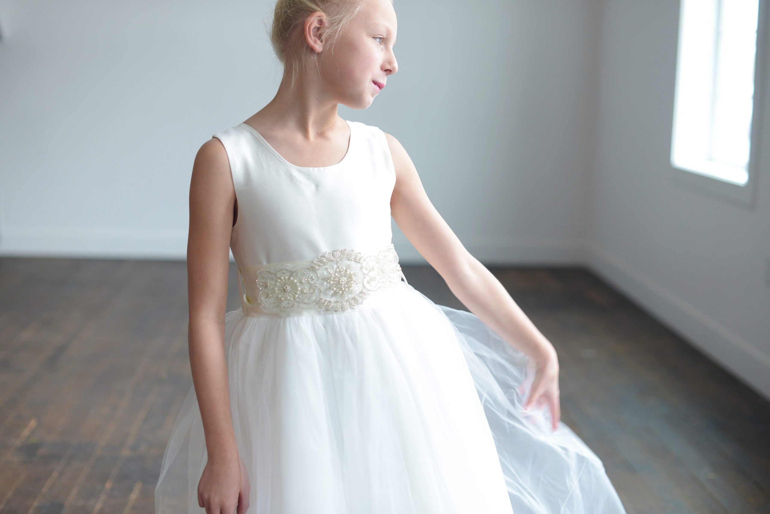 A photo of a white satin flower girl dress with a full ballerina style skirt in tulle and a diamante motif.