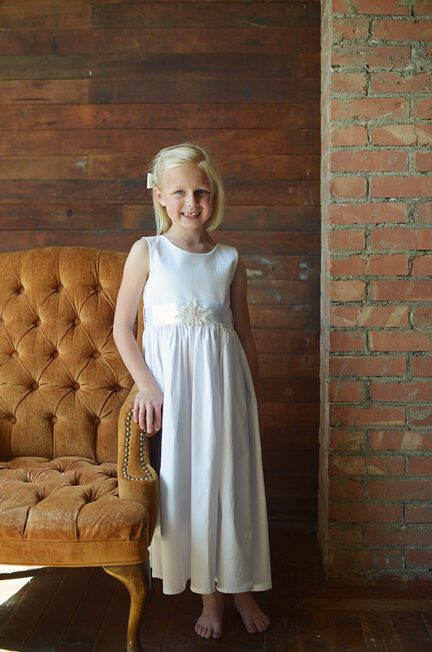 A photo of a girl wearing a summer flower girl dress in white cotton with a diamante sash