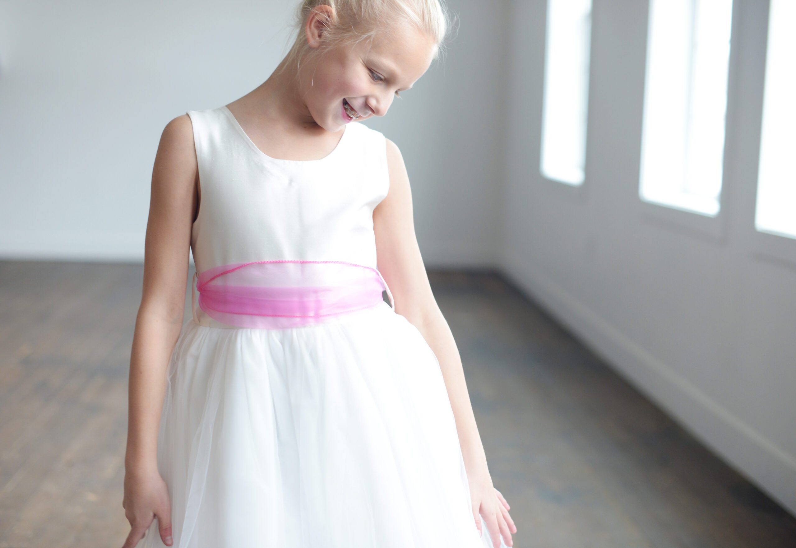 A photo of a girl wearing an ivory flower girl dress with a silk bodice and tulle skirt. The sash is a pink organza.