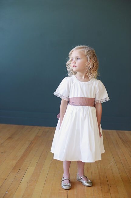 A photo of a flower girl wearing an ivory cotton flower girl dress with a lace trim on the neckline and an oversized dusky pink bow