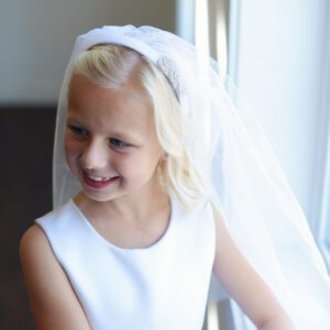 A photo of an 8 year old girl wearing a white satin first communion dress with an oversized bow and diamante and a veil