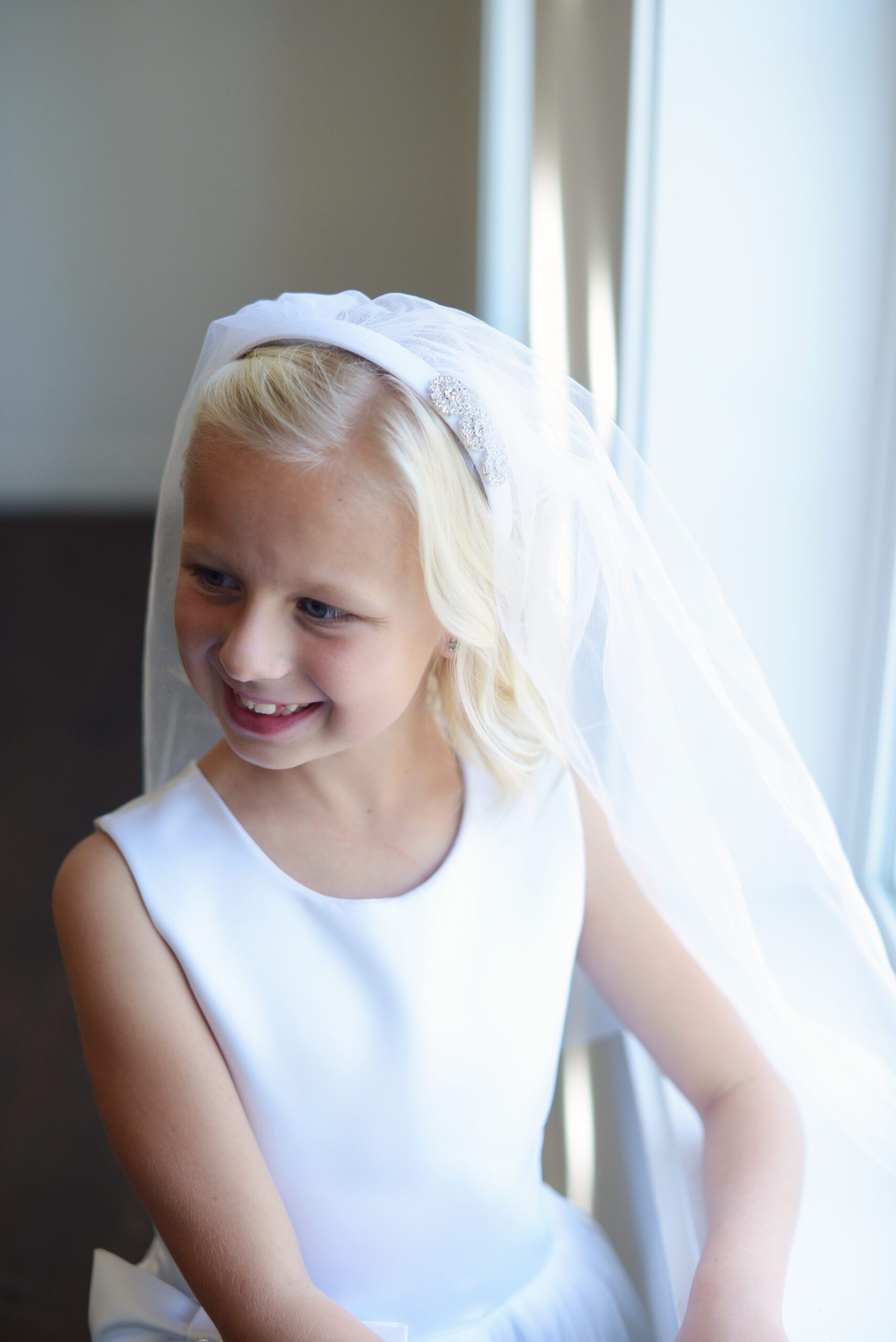 A photo of an 8 year old girl wearing a white satin first communion dress with an oversized bow and diamante and a veil