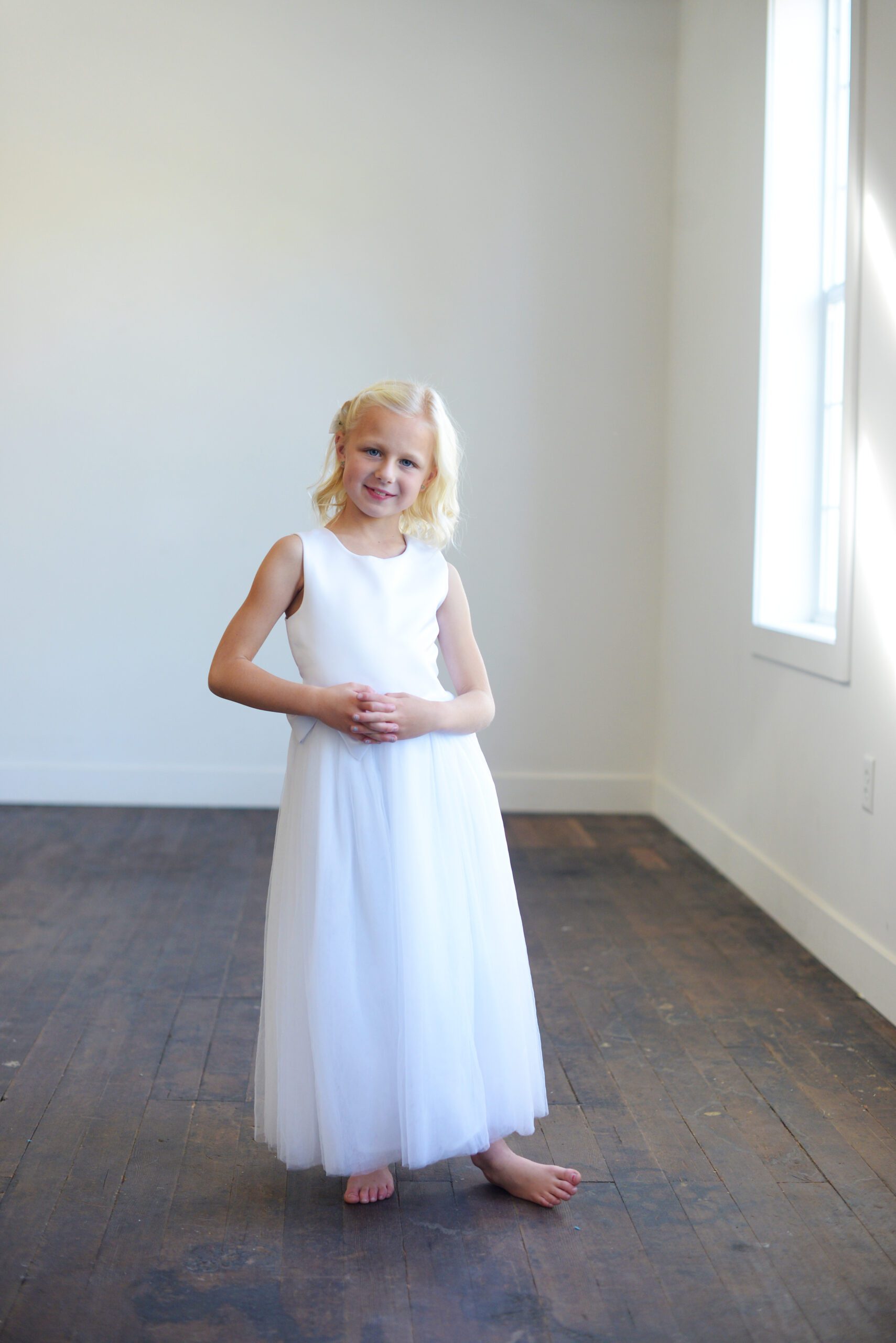 A photo of an 8 year old girl wearing a white satin first communion dress with an oversized bow and diamante
