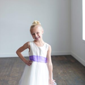 A photo of a girl wearing an ivory flwer girl dress with a silk bodice and tulle skirt. The sash is a purple organza.