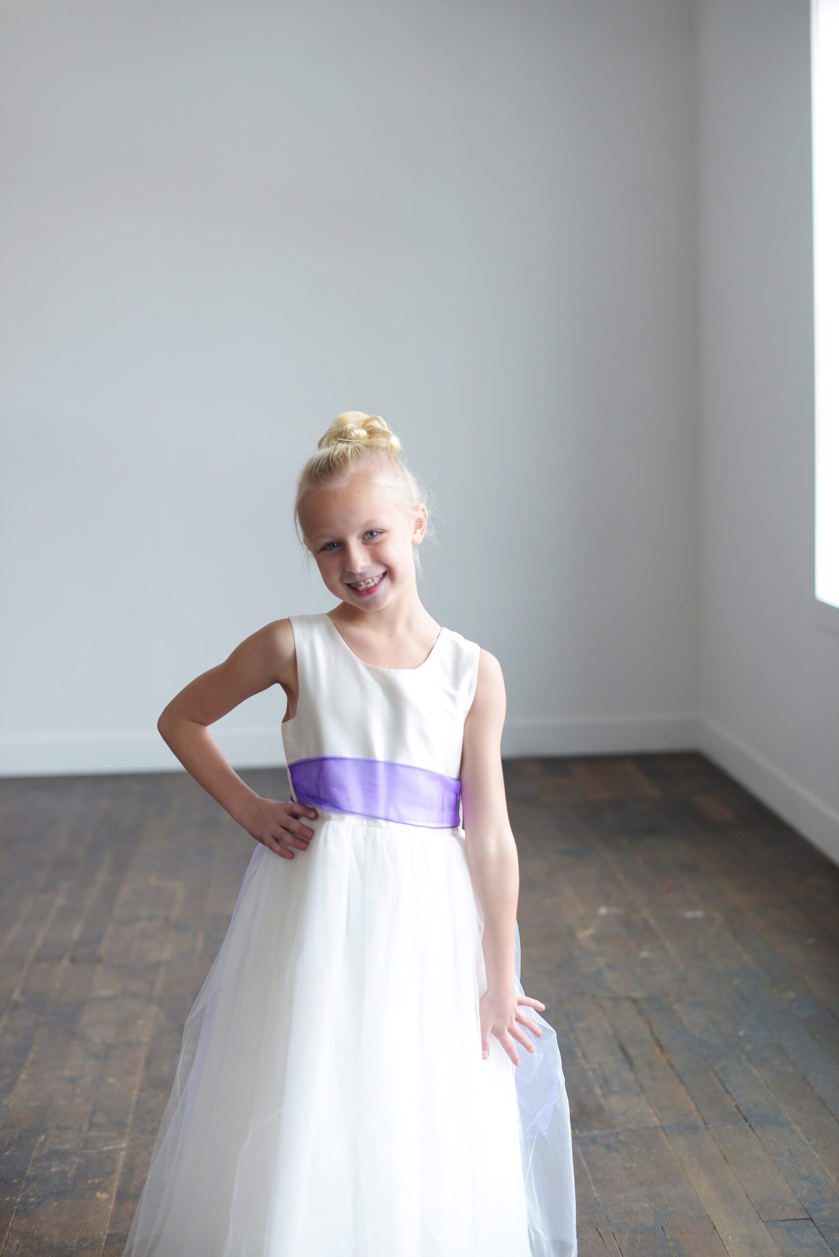 A photo of a girl wearing an ivory flwer girl dress with a silk bodice and tulle skirt. The sash is a purple organza.