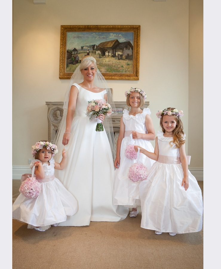 A group of flower girls wearing an ivory silk dress with a ruffle around the collar. The dress has a wide silk belt with an oversized bow
