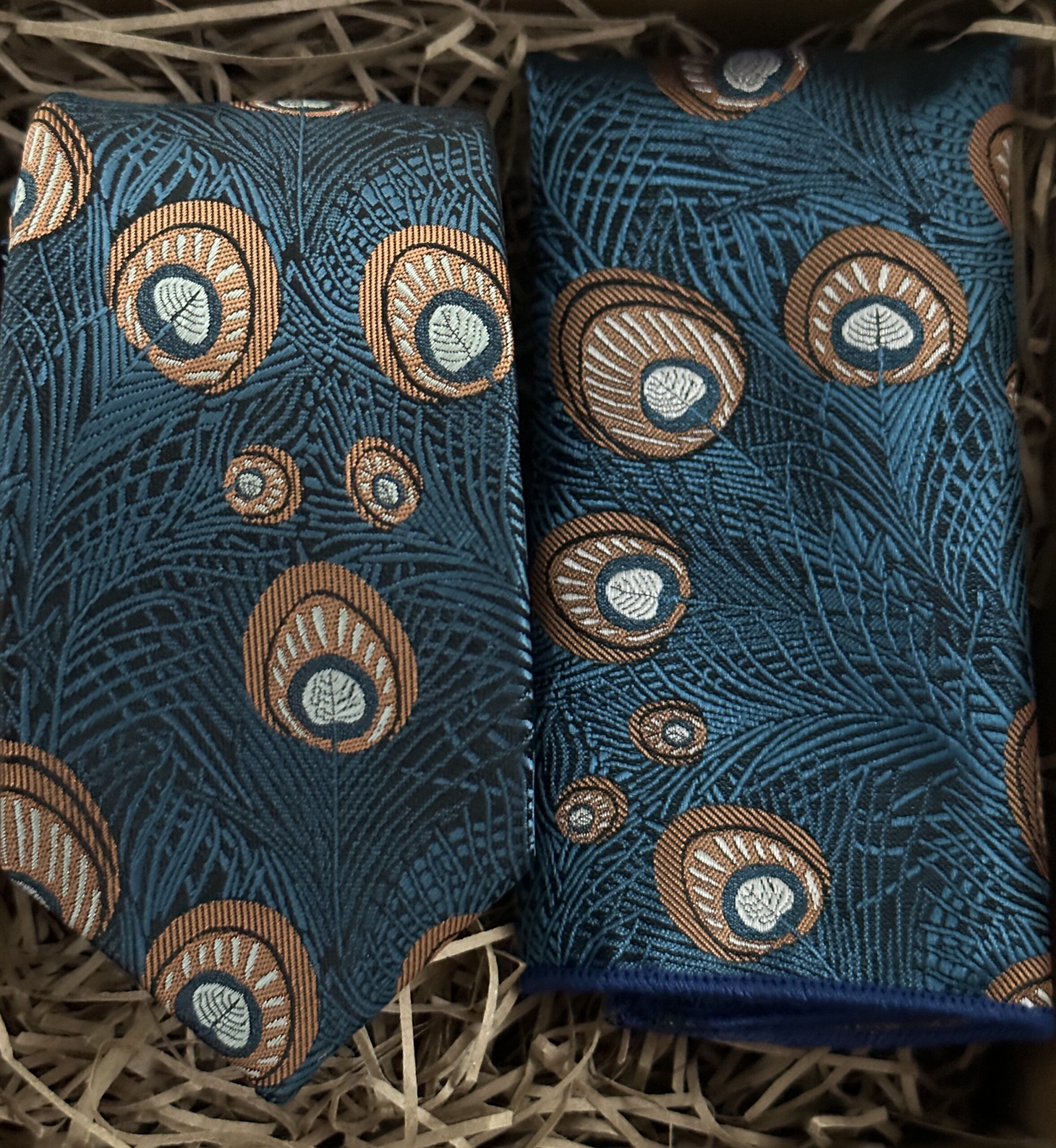 A photo of a peacock necktie and pocket square in a blue shade with gold feather plumes